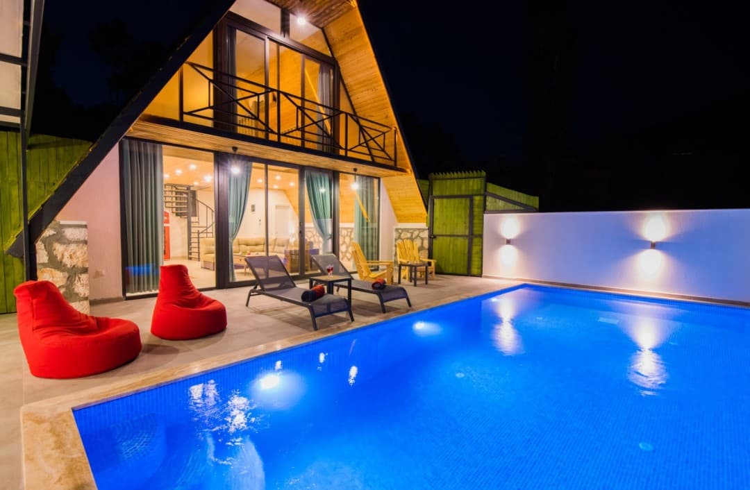 Gece aydınlatmalı havuz başında kırmızı minderli şezlonglar ve üçgen villa cephesi.