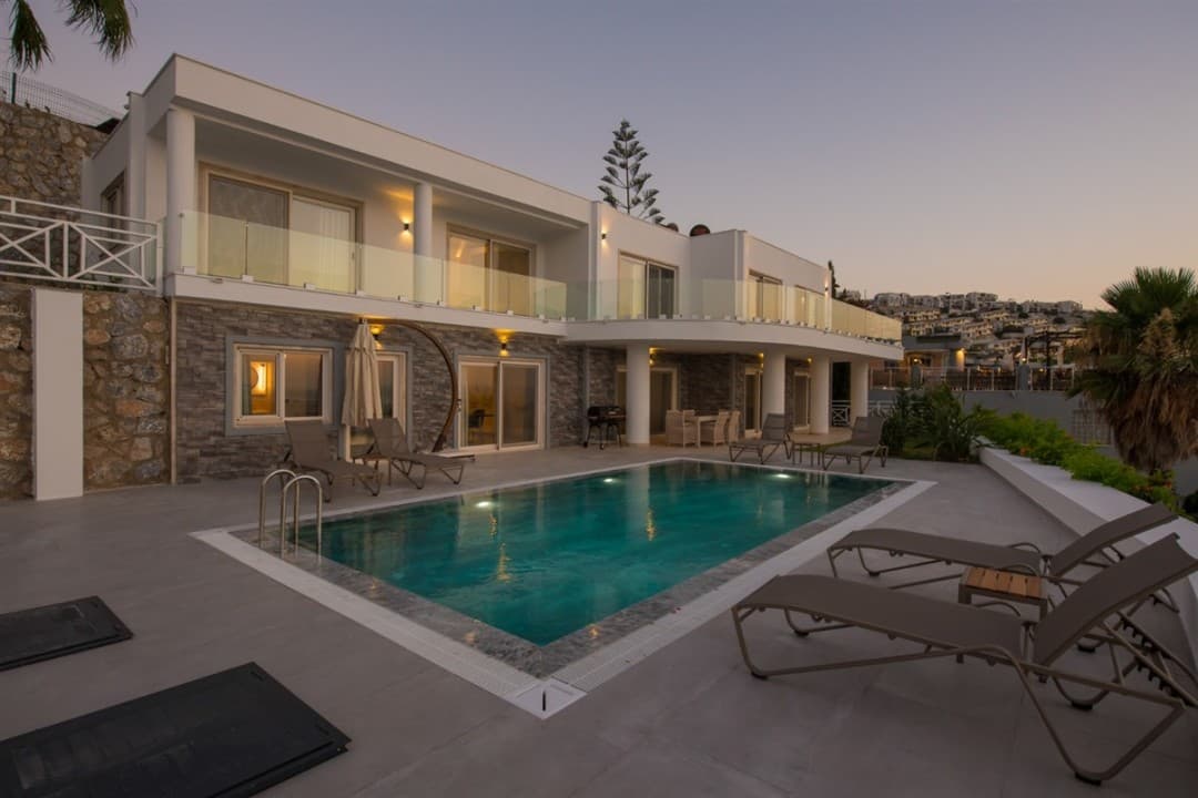 Akşam vakti aydınlatmalı havuzu ve şezlongları bulunan iki katlı modern villa.