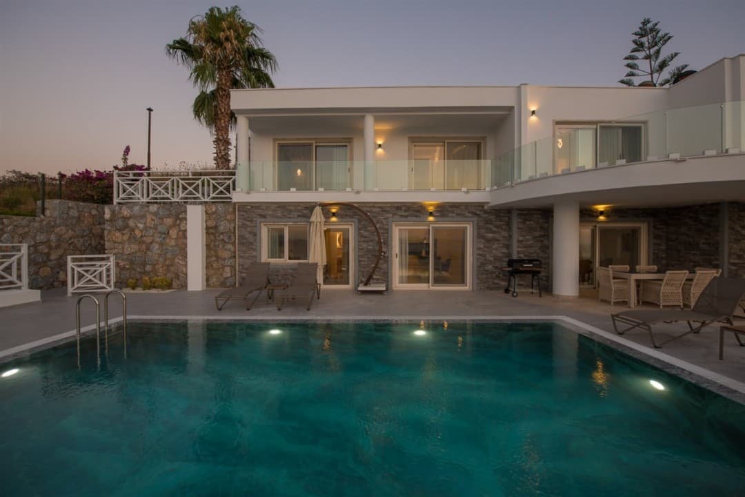 Akşam vakti aydınlatılmış havuzu ve şezlongları bulunan iki katlı modern villanın dış cephesi.