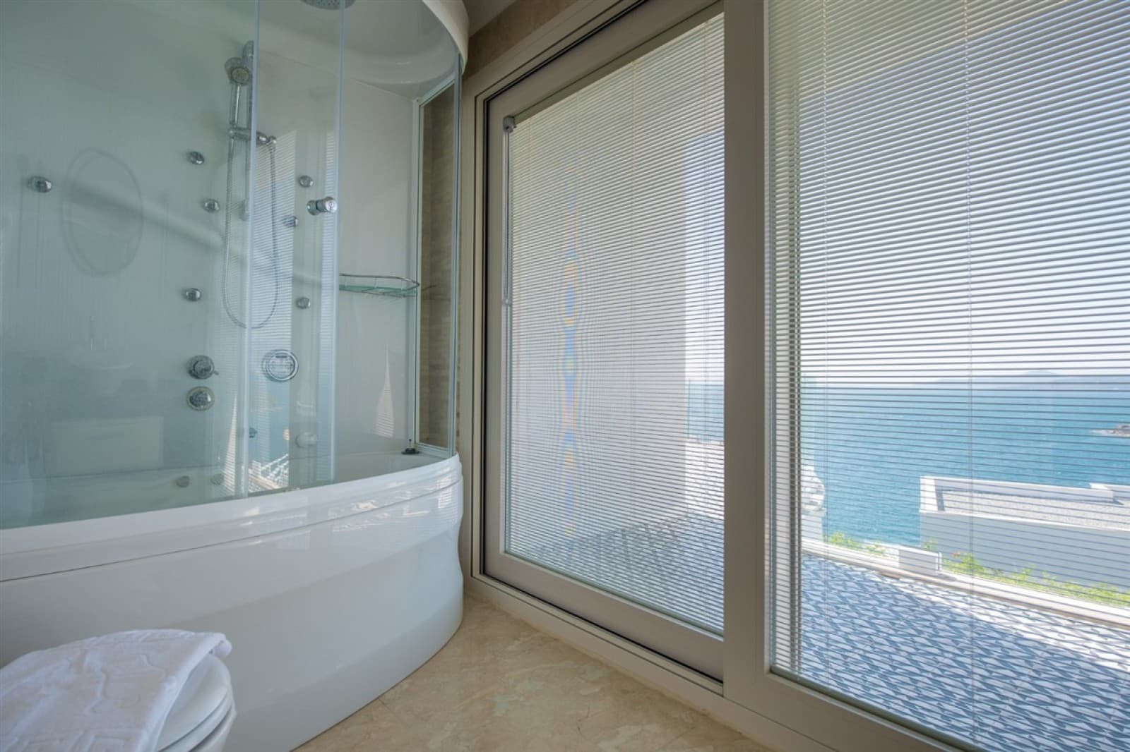 Deniz manzaralı büyük pencerelerin yanında yer alan beyaz küvetli ve duşakabinli banyo.