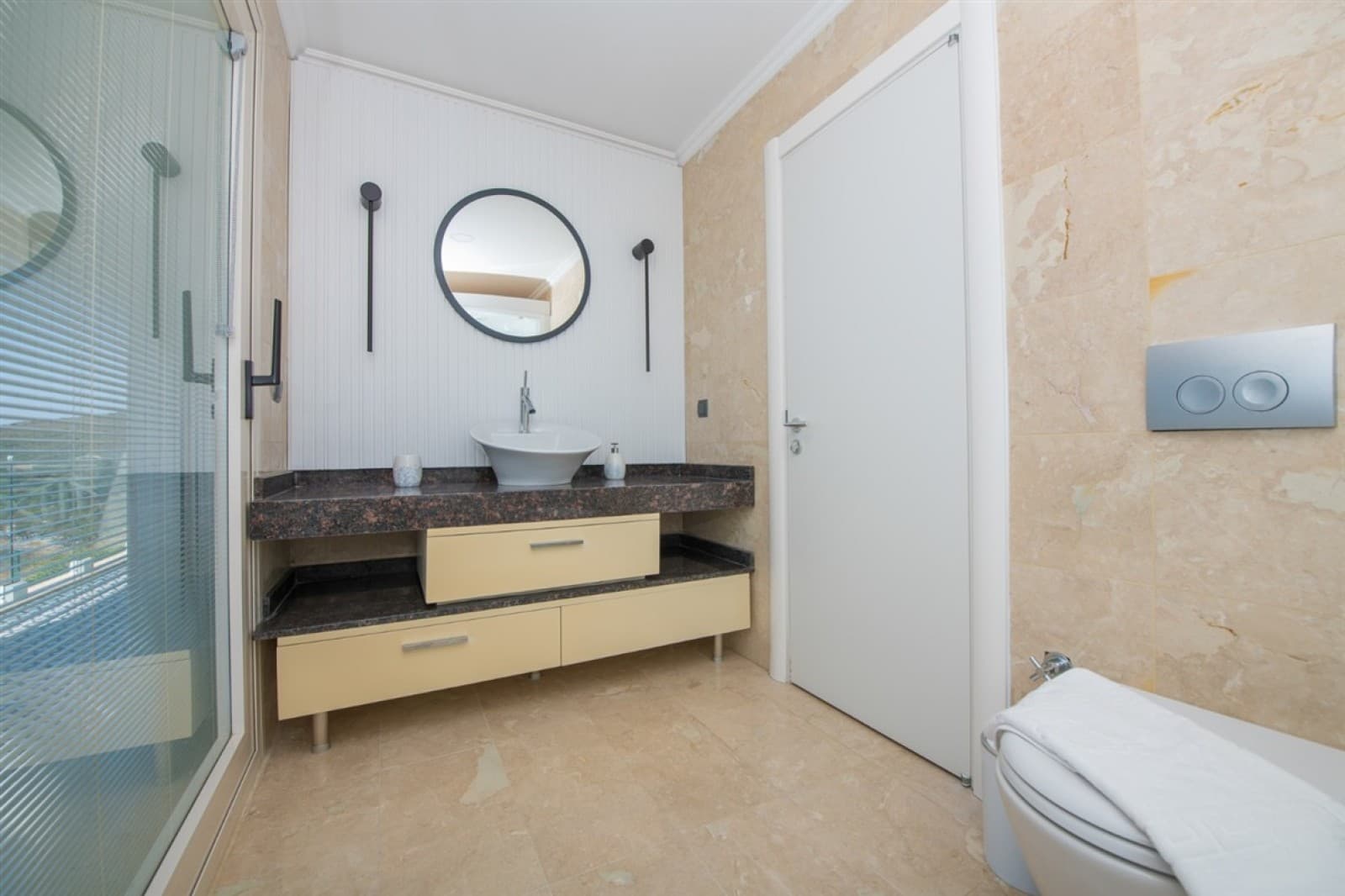 Bej mermer kaplı duşakabin, yuvarlak aynalı lavabo ve klozetli banyo.