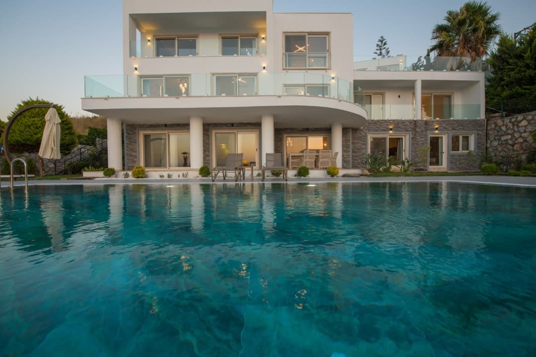 Önünde havuz bulunan, balkonlu ve cam korkuluklu iki katlı beyaz modern villa.