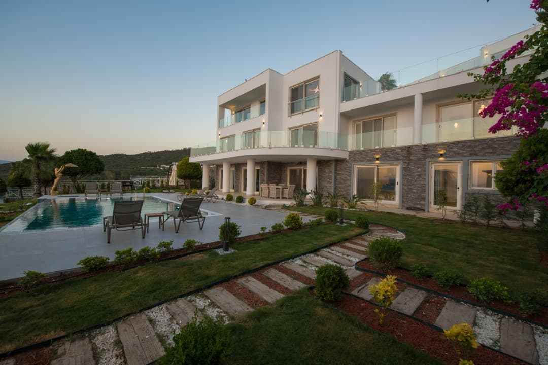 Önünde yüzme havuzu ve şezlonglar bulunan, taş kaplamalı zemin katı ve beyaz dış cephesi olan üç katlı villa.