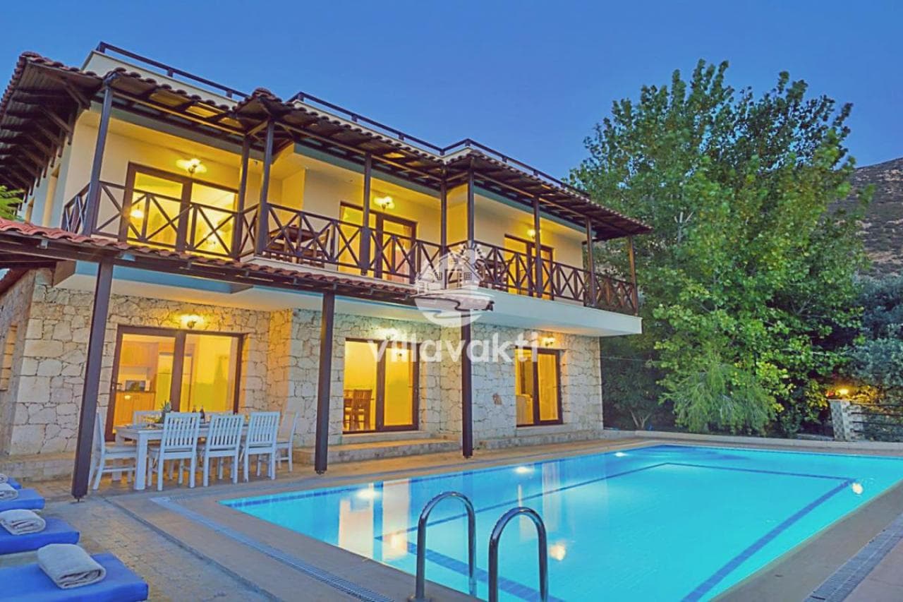 Gece aydınlatmalı, ahşap balkonlu iki katlı taş villa ve önünde yüzme havuzu.