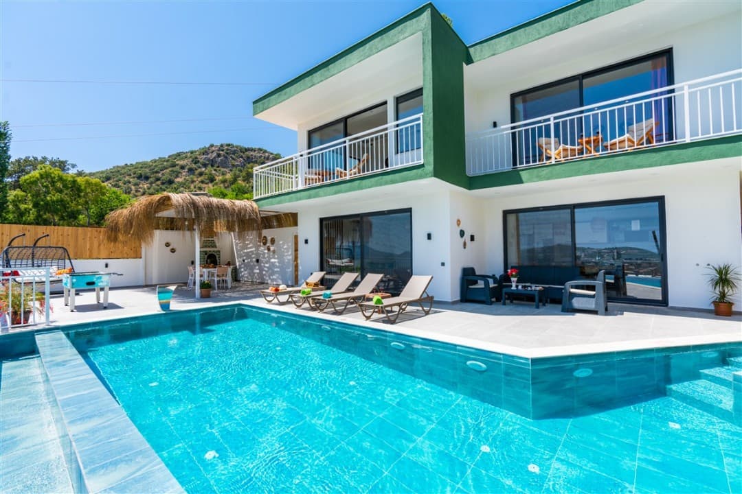 Önünde güneşlenme şezlongları ve havuz bulunan, yeşil beyaz dış cepheli iki katlı modern villa.