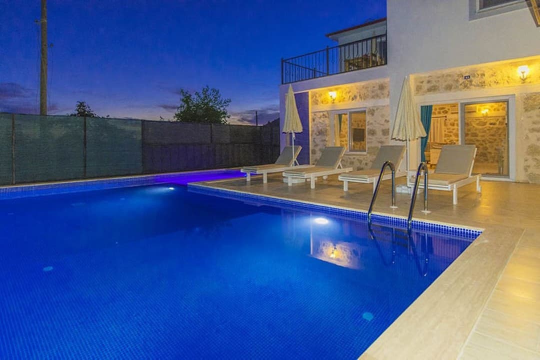 Akşam vakti havuz kenarında şezlonglar ve iki şemsiye bulunan iki katlı villa.
