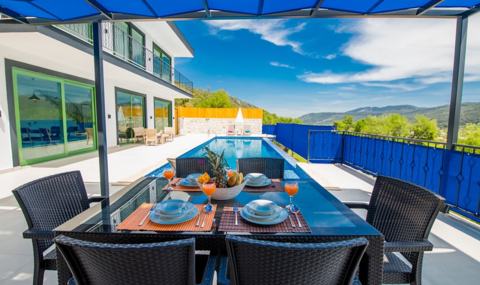 Mavi tenteli terasta yemek masası, arkada havuz ve dağ manzaralı villa.