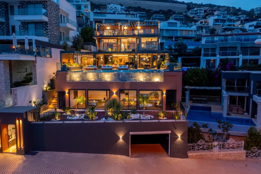 Akşam ışıklarıyla aydınlatılmış, sonsuzluk havuzlu ve teraslı modern çok katlı villa dış görünüşü.