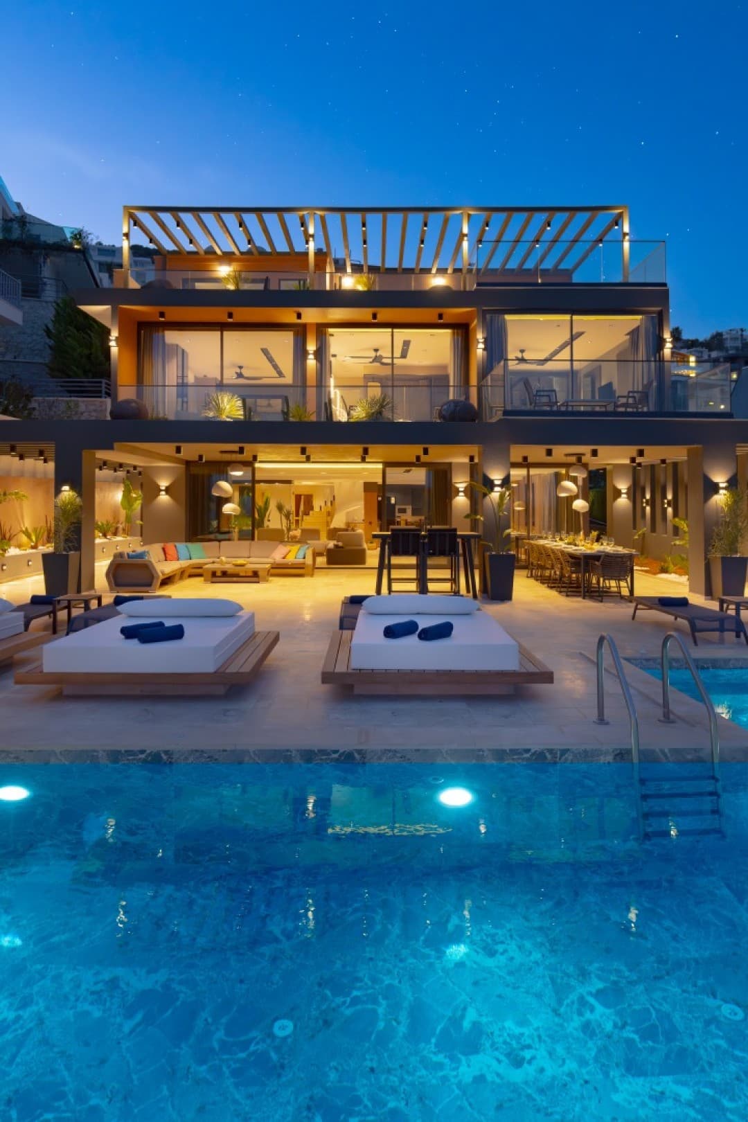 Akşam vakti havuz başı şezlongları görünen aydınlatılmış modern villa.