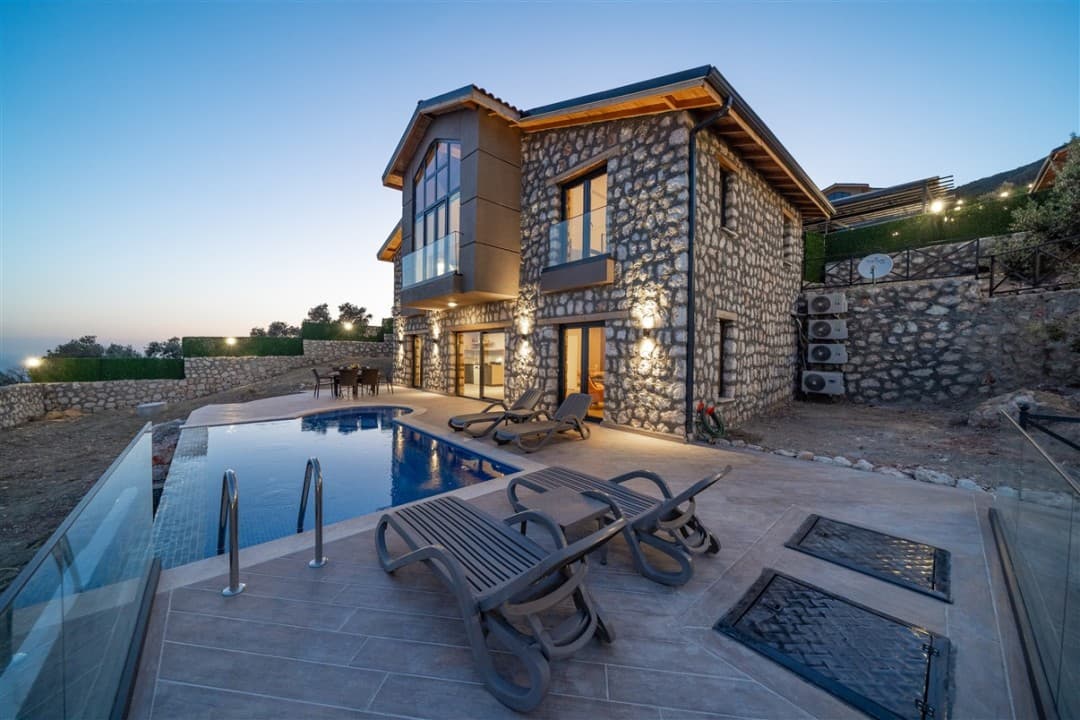 Akşam vakti aydınlatılmış taş kaplama iki katlı villa, özel yüzme havuzu ve şezlonglar.