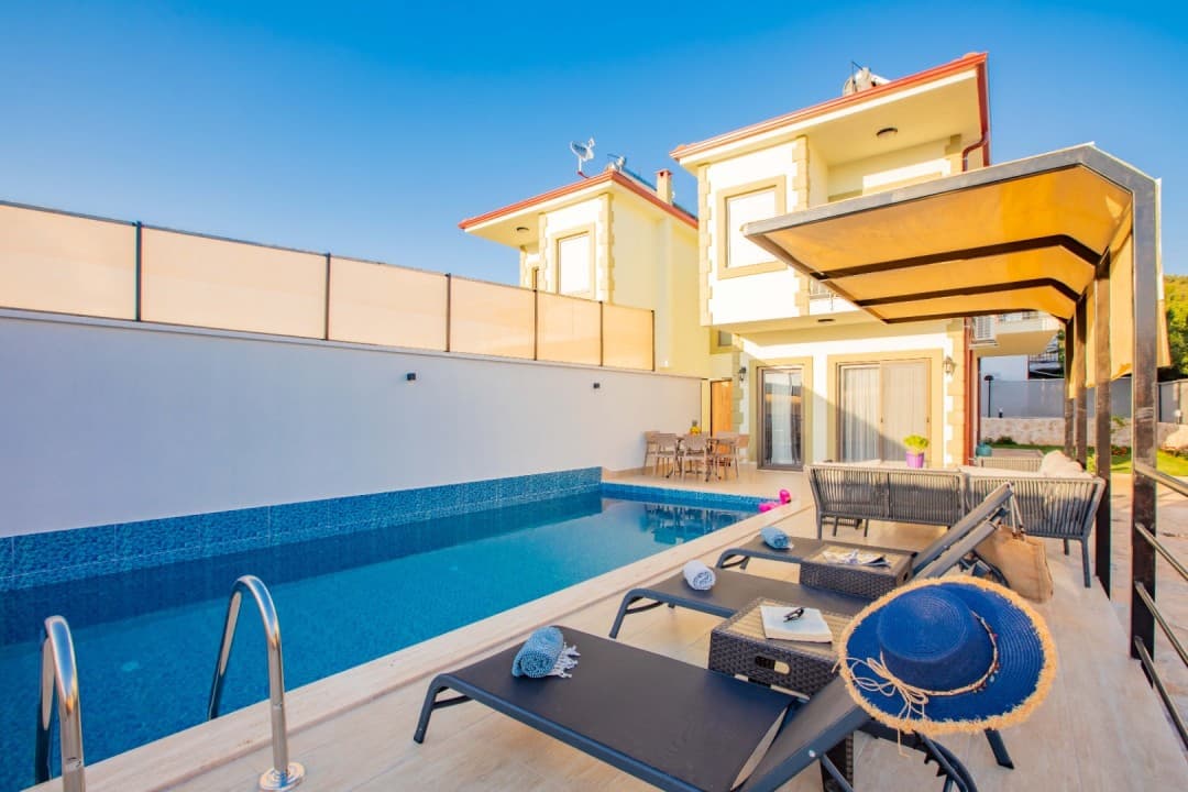Güneşli günde havuz başı şezlongları ve arkasında iki katlı bej villa.