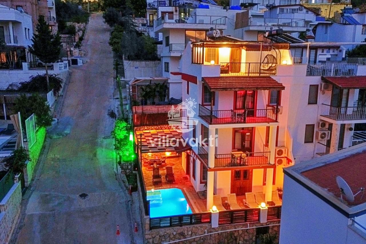 Akşam aydınlatması altında üç katlı villa, özel yüzme havuzu ve sokak görünümü.