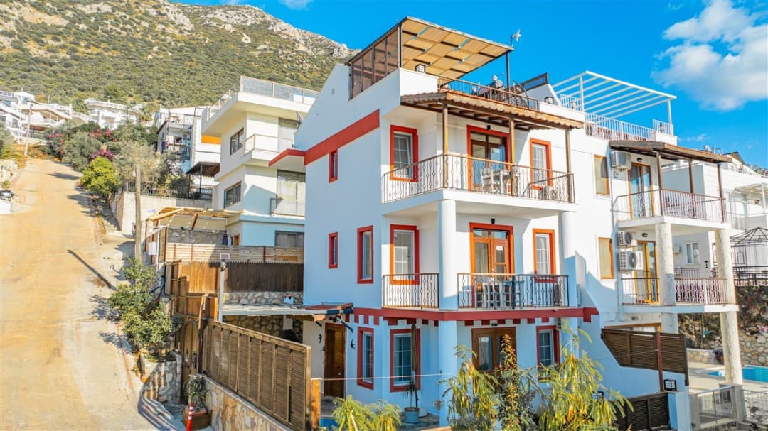 Beyaz cepheli, kırmızı çerçeveli pencereleri olan üç katlı villa ve yokuş sokak.