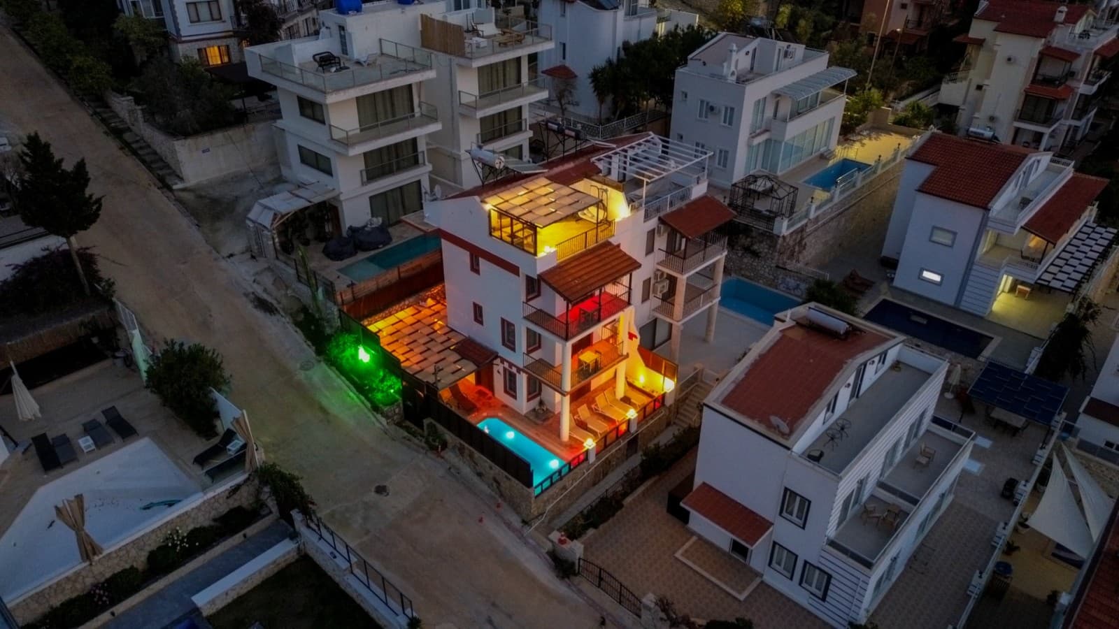 Gece çekiminde aydınlatılmış müstakil villanın havuzlu terası ve çevre binalar.