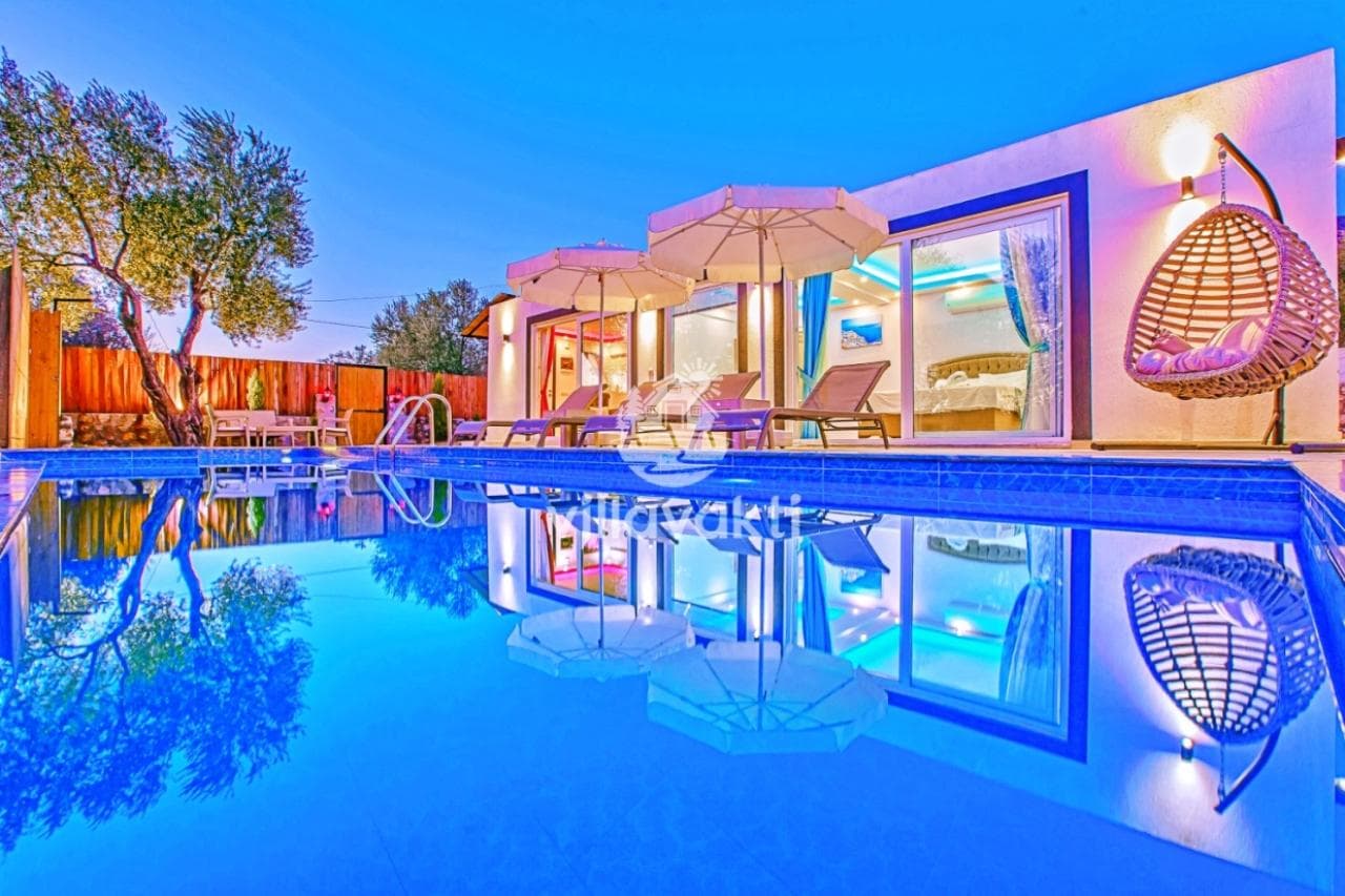 Gece aydınlatmalı yüzme havuzuna yansıyan villa, şezlonglar ve salıncak.