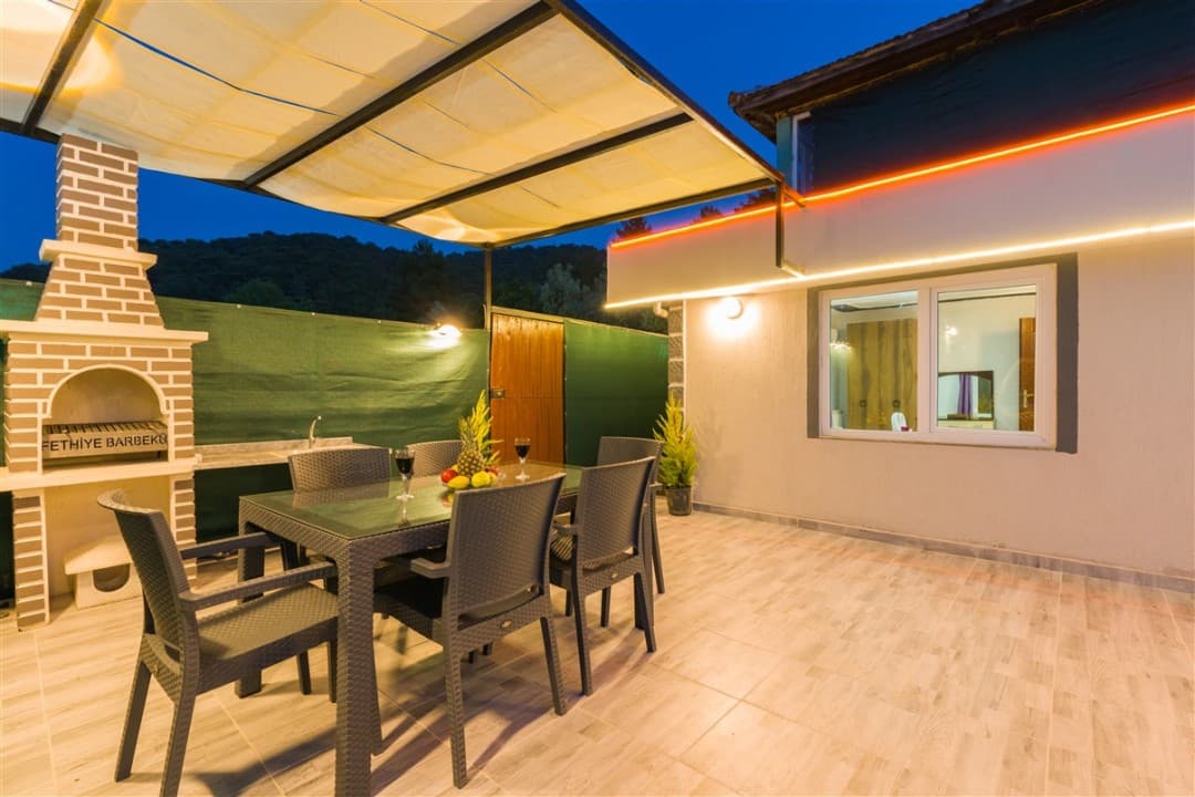 Akşam vakti aydınlatmalı verandasındaki yemek masası ve tuğla barbekü bulunan villa bahçesi.