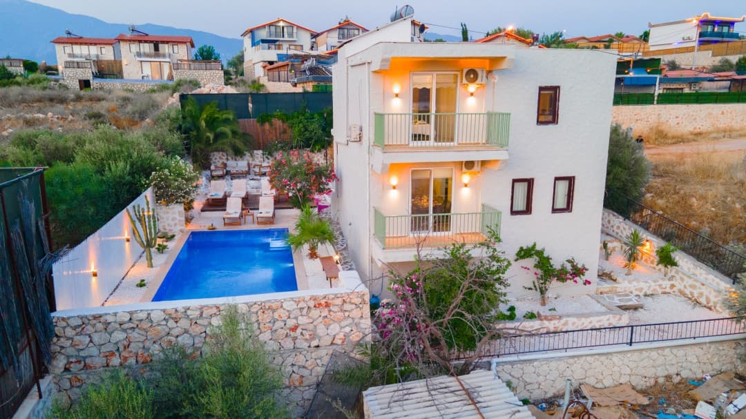 İki katlı beyaz villanın önünde havuz, şezlonglar ve dağ manzarası var.