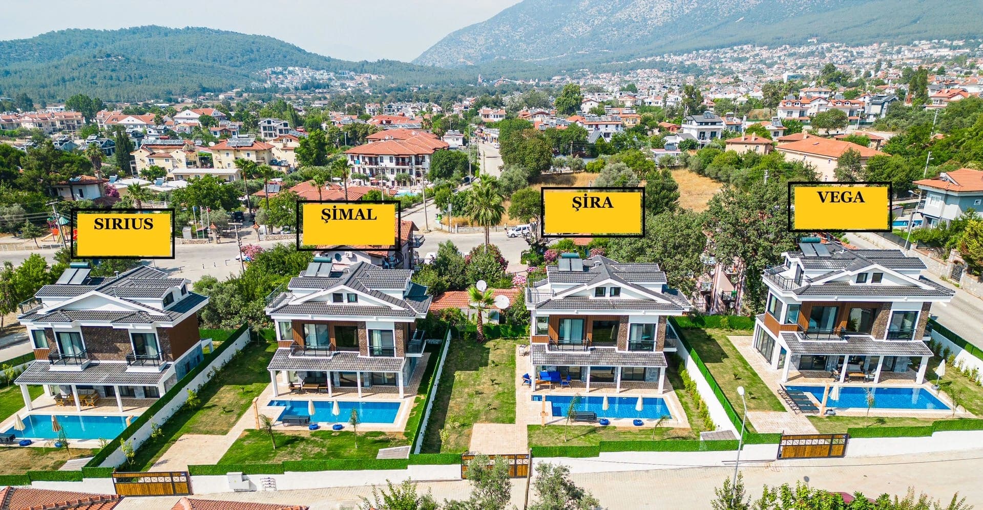 Dağ ve şehir manzaralı bir alanda yer alan havuzlu villaların tepeden görünümü.