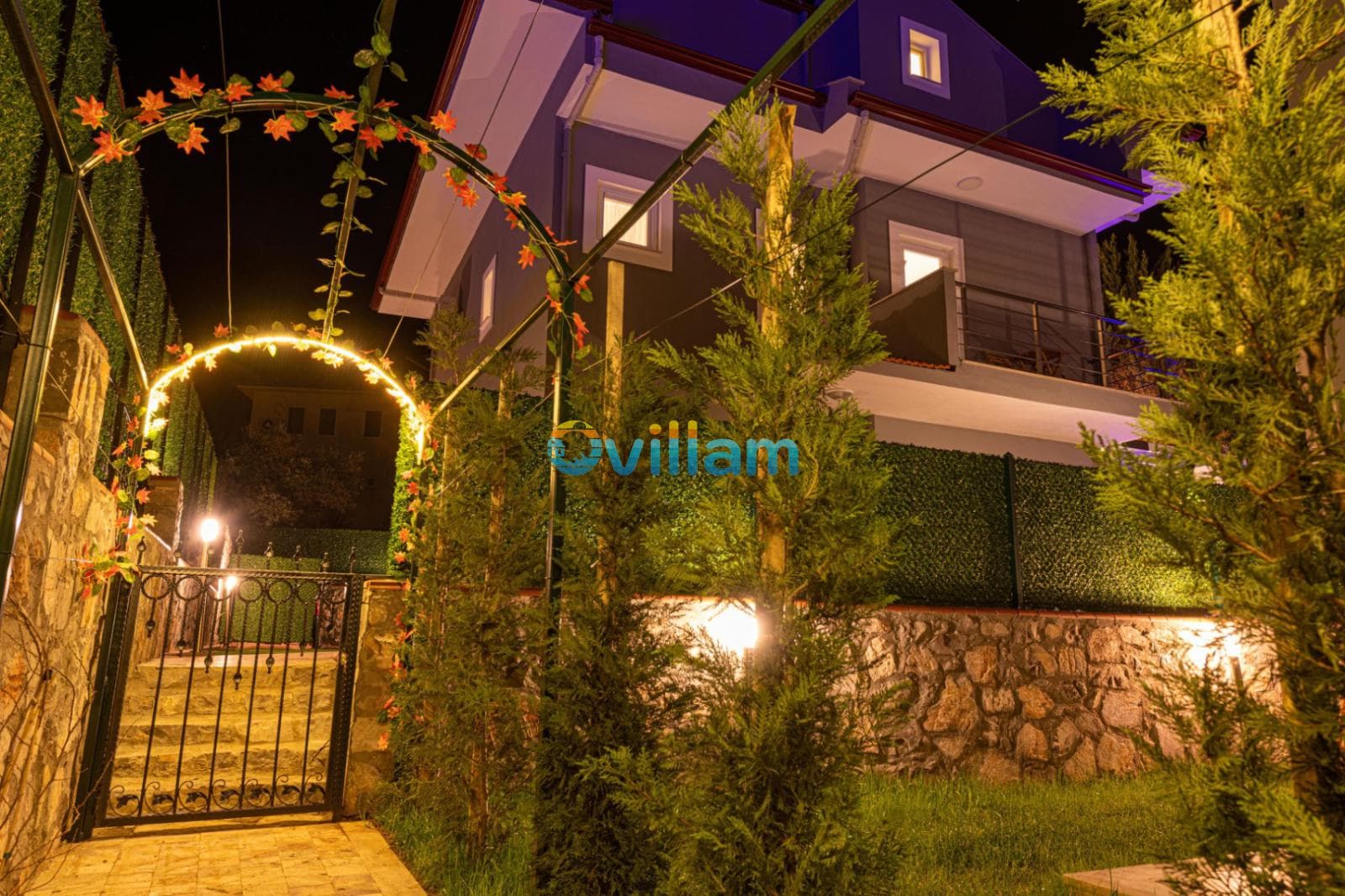 Gece aydınlatılmış demir bahçe kapısı, ışıklı bitki takı ve arkasında villa binası.
