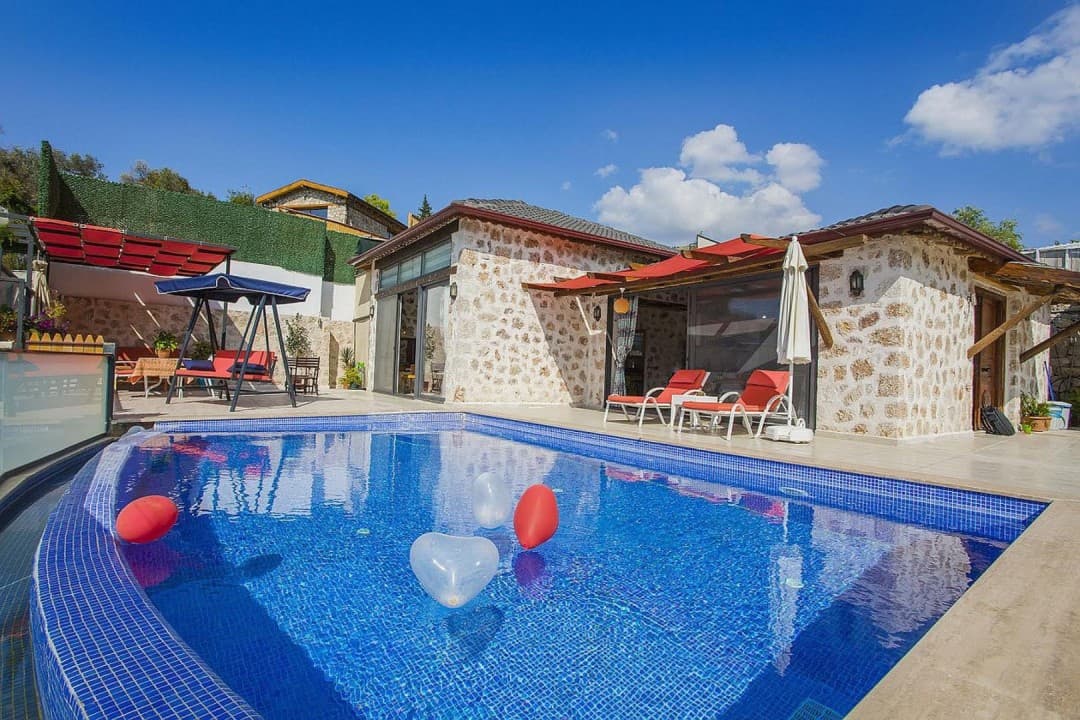 Taş duvarlı villa, önünde havuz, havuz içinde balonlar, şezlonglar ve bahçede salıncak bulunan güneşli alan.