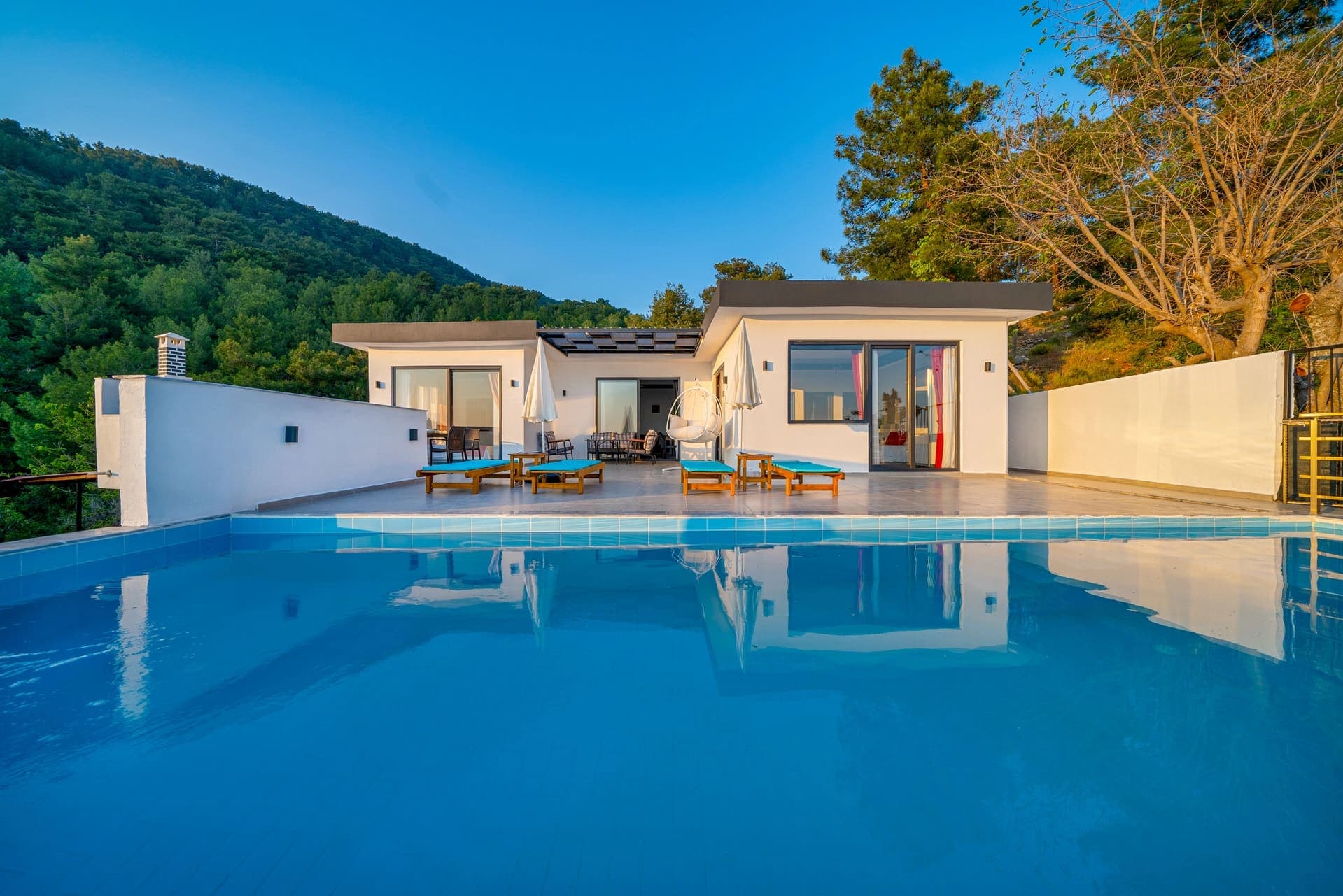 Geniş yüzme havuzunun arkasında şezlongların yer aldığı tek katlı villa ve dağ manzarası.