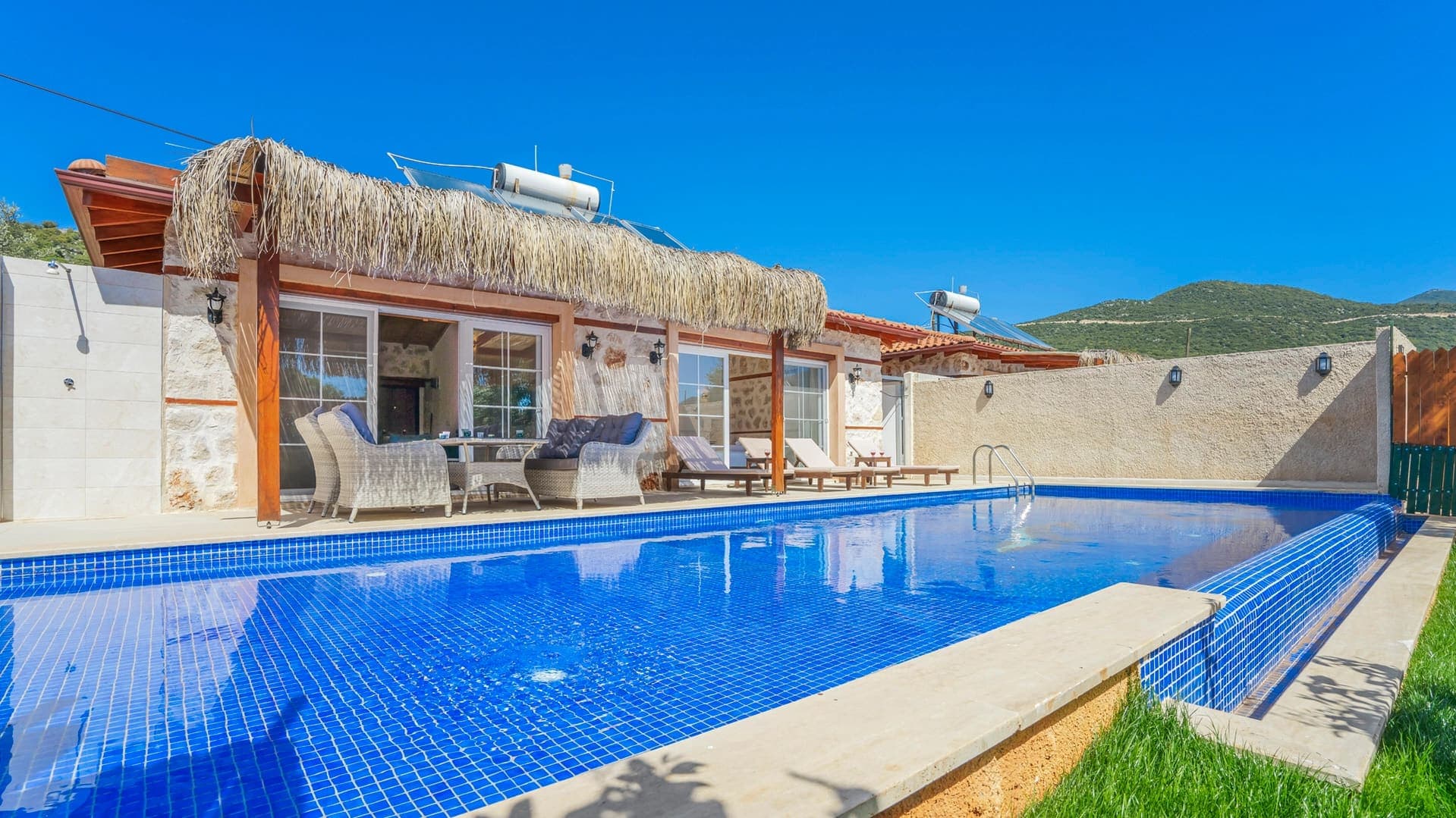 Mavi fayanslı yüzme havuzu, şezlonglar ve saz çatılı verandaya sahip taş villa.