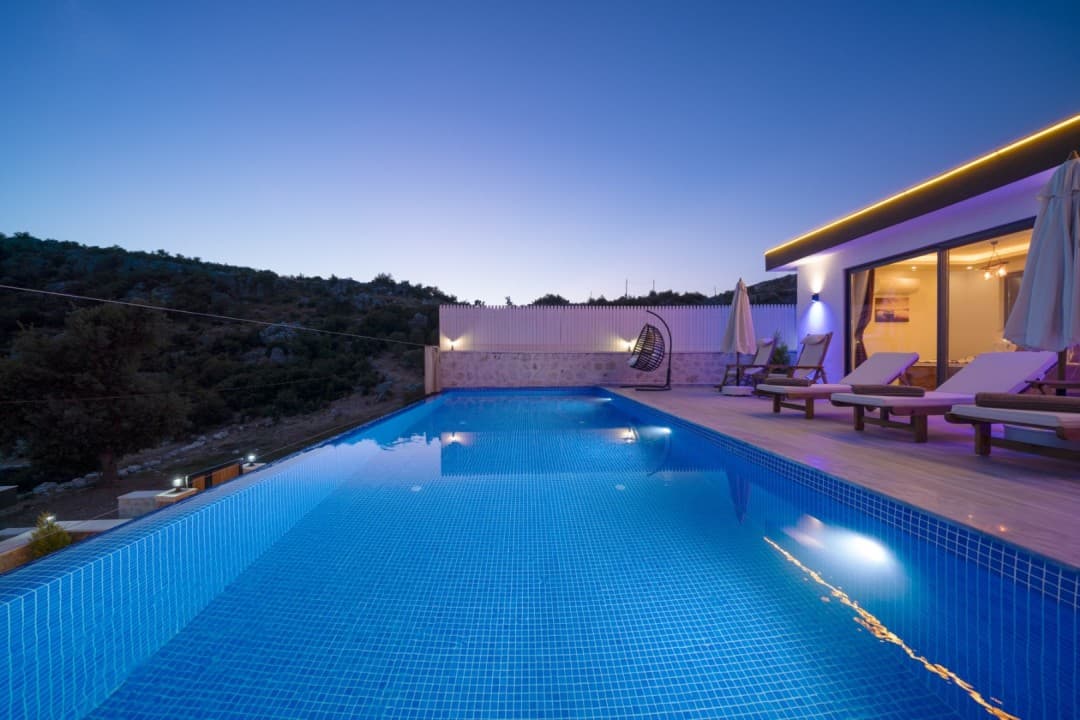 Gün batımında sonsuzluk havuzu, şezlonglar ve arka planda tepelik arazi bulunan modern villa terası.