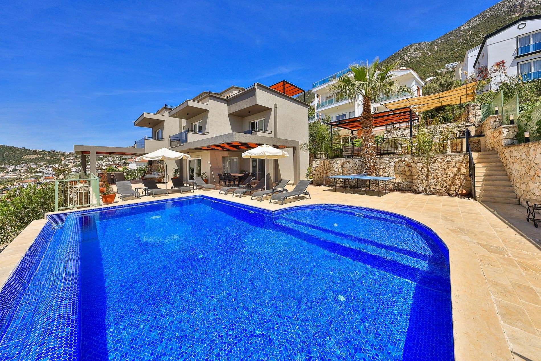 Geniş yüzme havuzu, şezlonglar, masa tenisi ve arkada modern iki katlı villa.
