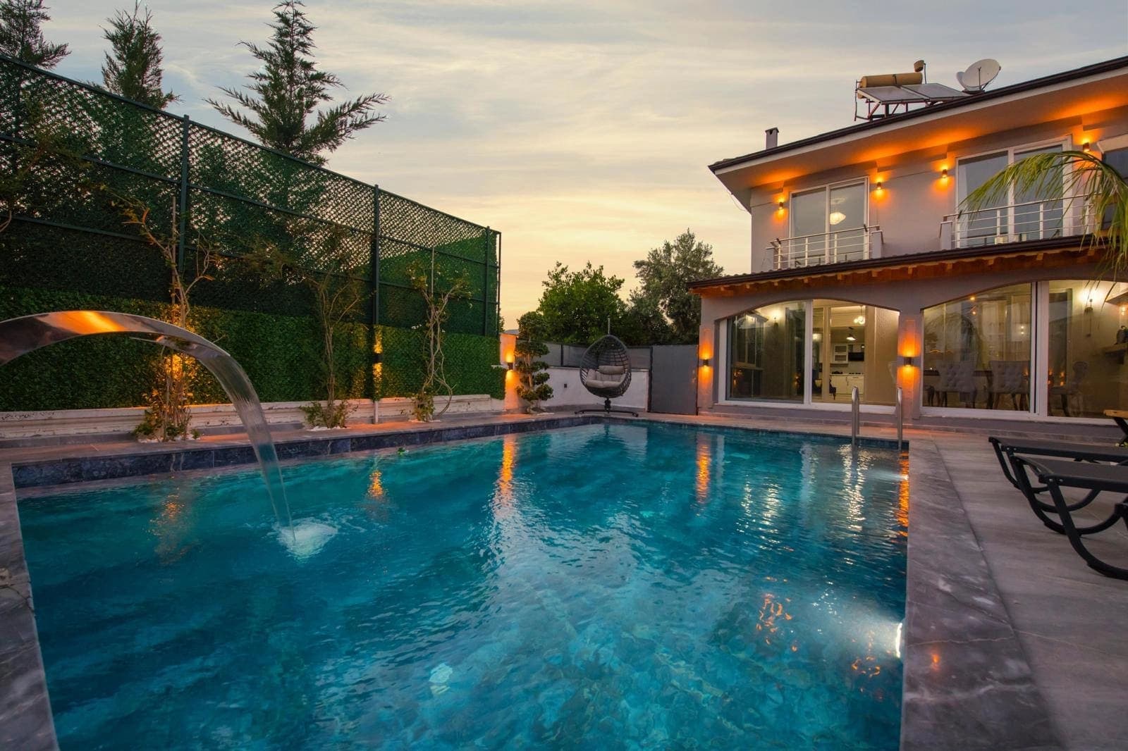 Akşamüstü aydınlatmalı havuz, şezlonglar ve iki katlı villa.