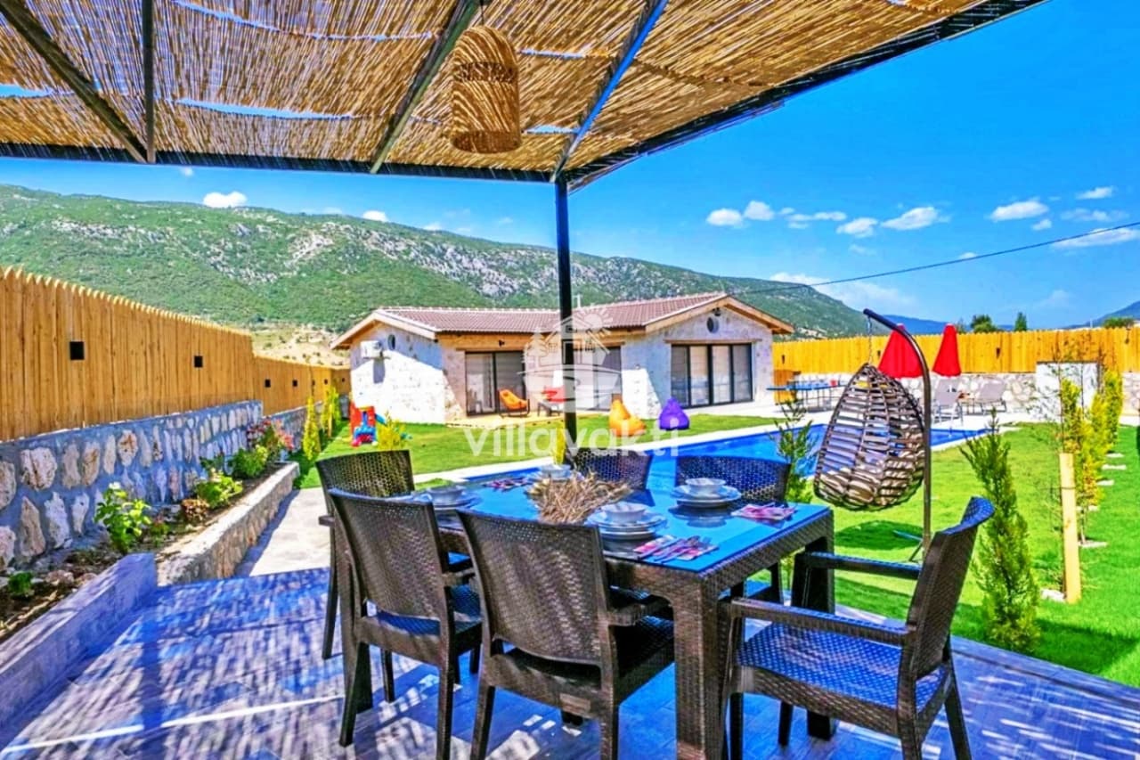 Hasır gölgelikli veranda altında yemek masası, havuz ve dağ manzarası.