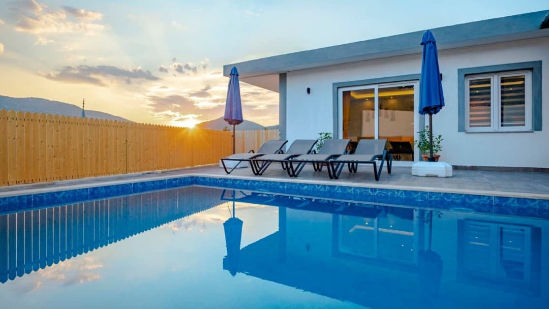 Gün batımında havuz başında şezlonglar ve iki mavi şemsiye bulunan villa.