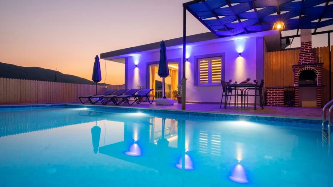 Gün batımında aydınlatmalı havuzu, şezlongları ve dış mekan yemek masası bulunan tek katlı villa.