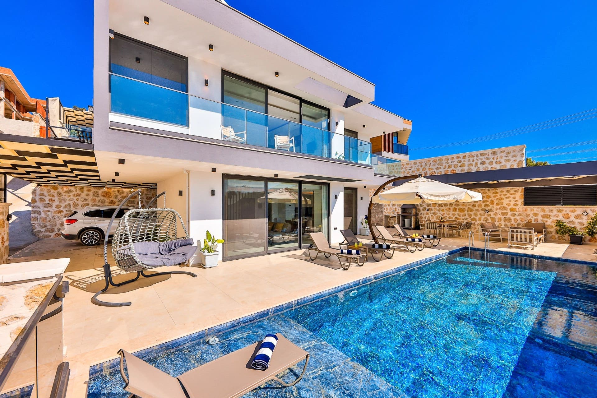 Önünde havuz ve şezlonglar bulunan, taş detaylı iki katlı modern villa.