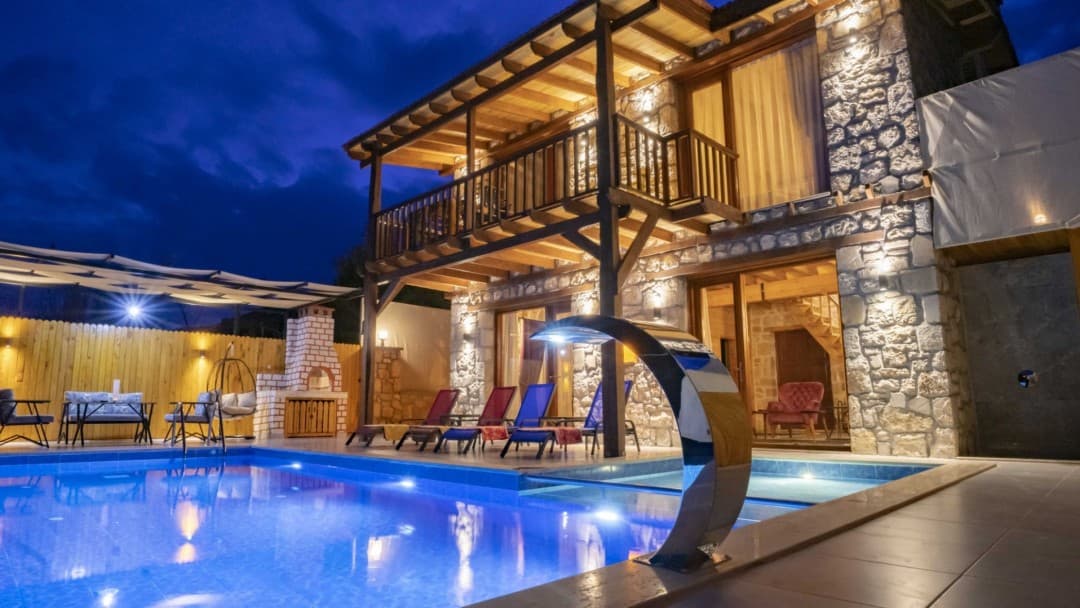 Akşam saatlerinde aydınlatmalı havuzu ve şezlongları olan iki katlı taş villa.
