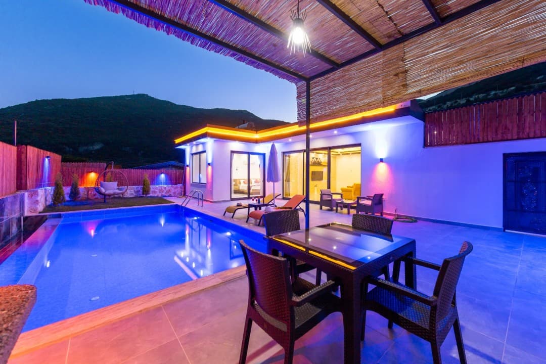 Akşam saatinde havuz kenarında masa, sandalyeler ve arka planda dağ manzaralı villa.