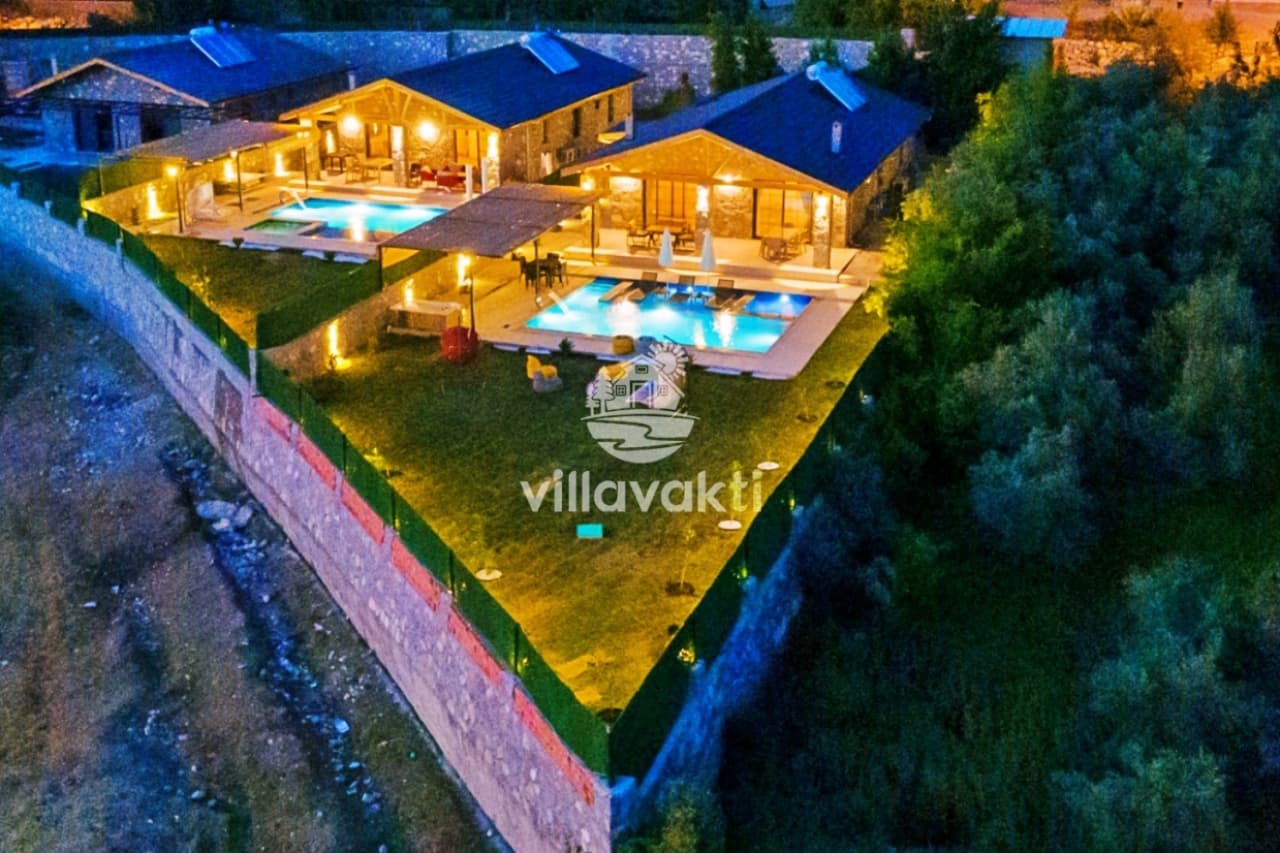 Aydınlatmalı havuzları ve yeşil bahçesi olan villaların akşam havadan görünümü.