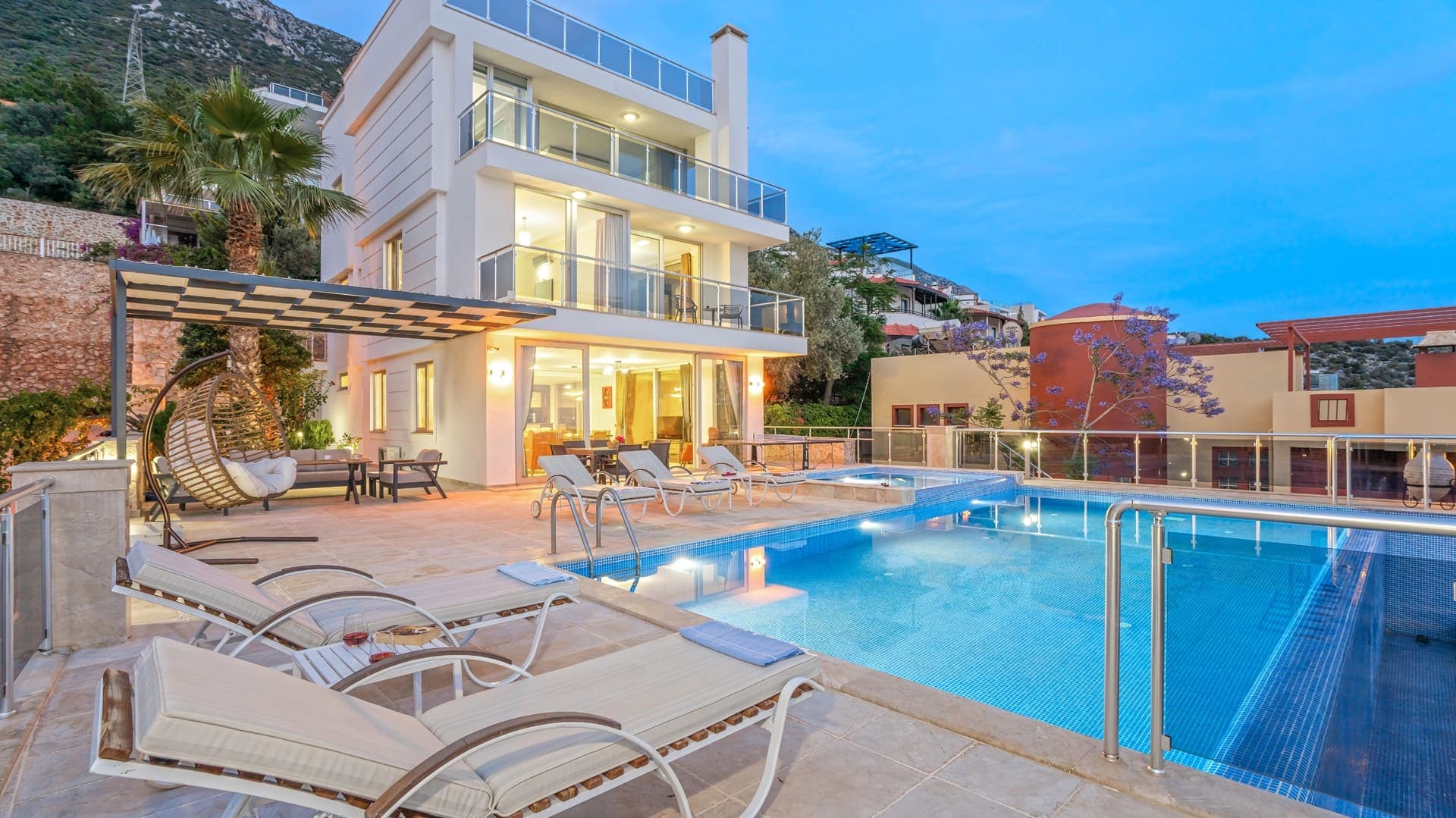 Önünde yüzme havuzu, şezlonglar ve salıncak bulunan beyaz dış cepheli villa.