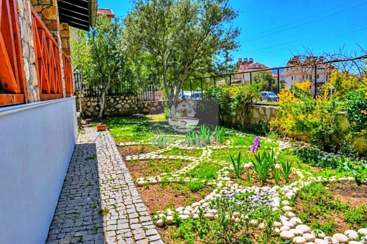 Taş duvarlarla çevrili yeşil bahçede taş döşeli yürüyüş yolu ve çiçek tarhları yer alıyor.