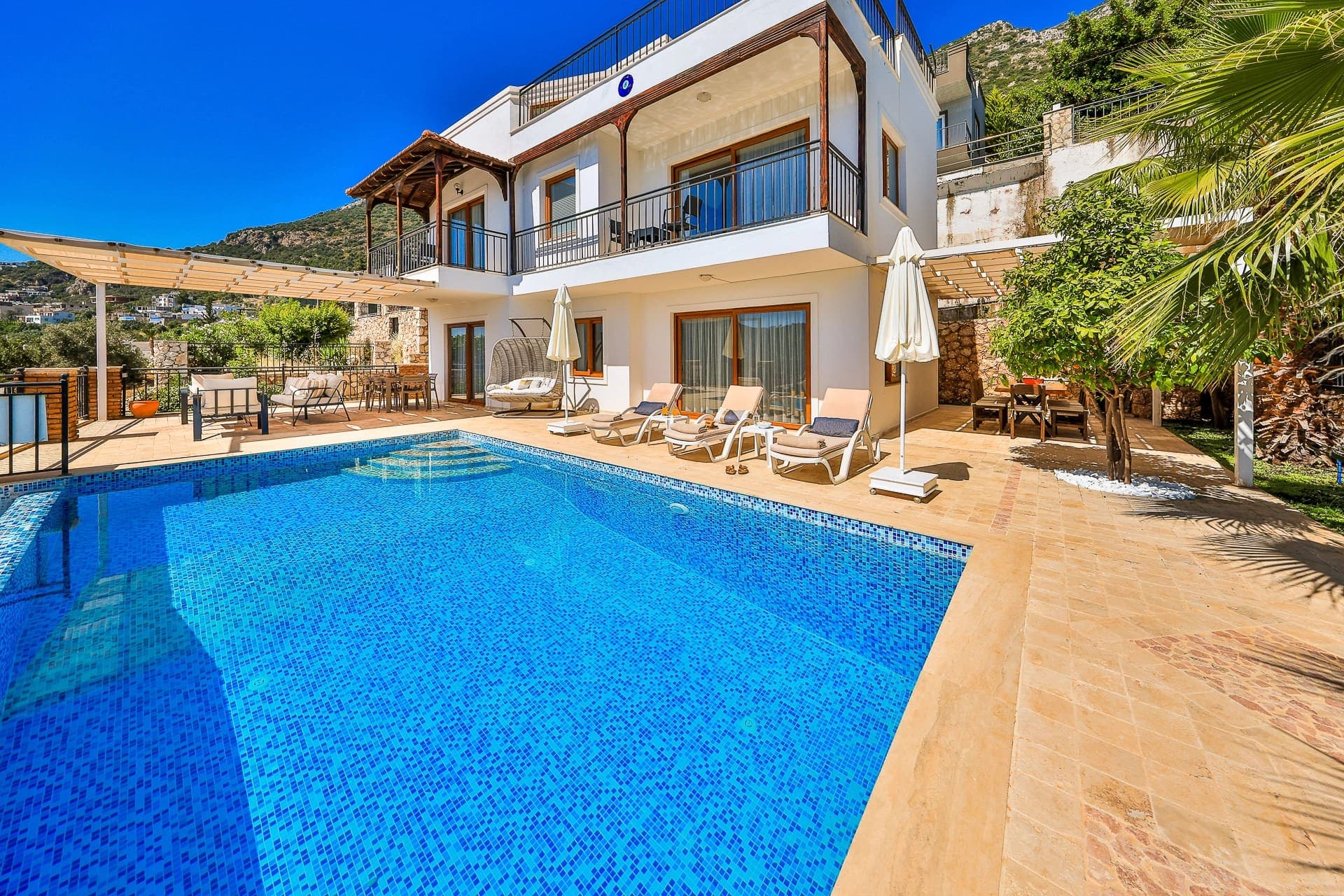 Balkonlu beyaz villa, havuz ve şezlongların bulunduğu güneşli dış cephe görünümü.