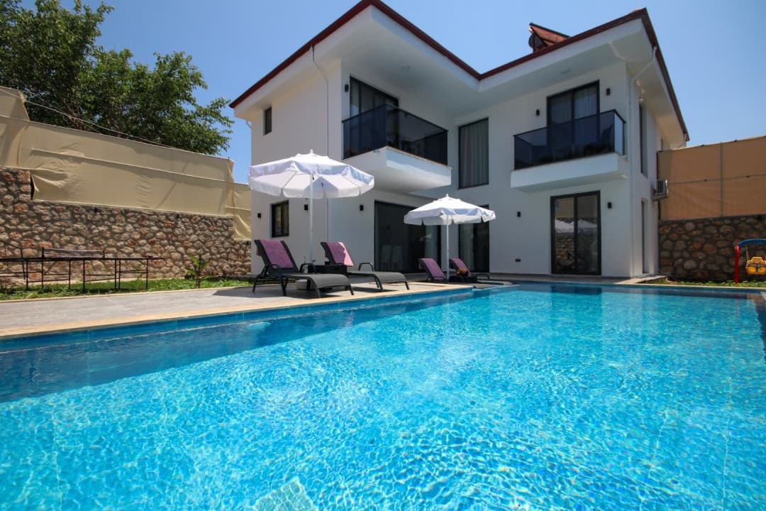 Güneşli günde havuz başında şezlonglar ve iki katlı beyaz villa.
