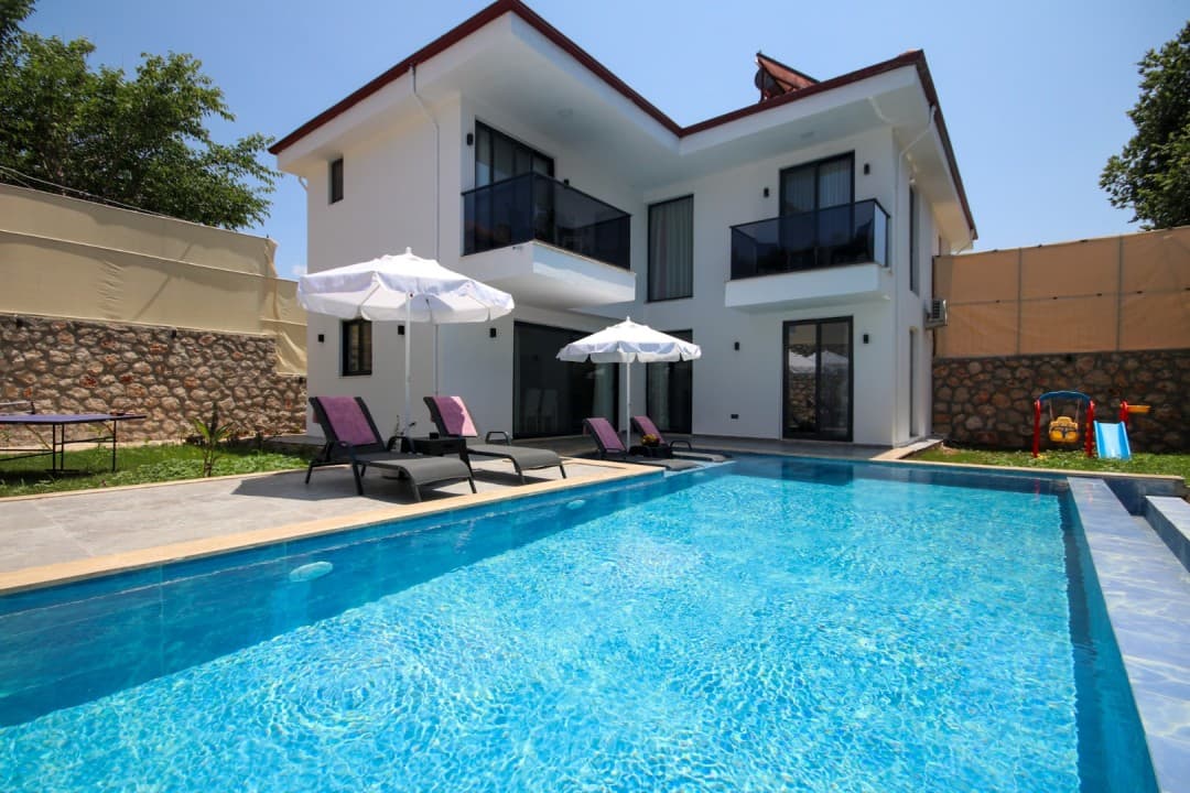 Önünde yüzme havuzu, şezlonglar ve güneş şemsiyeleri bulunan iki katlı beyaz dış cepheli villa.