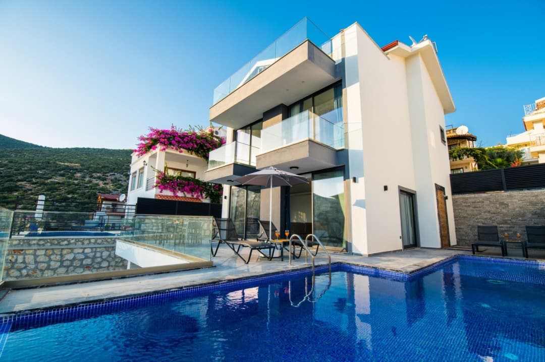 Mavi havuzlu, cam balkonlu modern villa ve havuz kenarında şezlonglar.