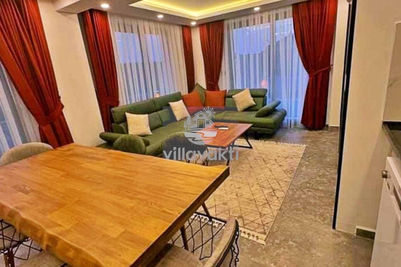 Ahşap yemek masası, yeşil köşe koltuk ve desenli halının bulunduğu iç mekan.
