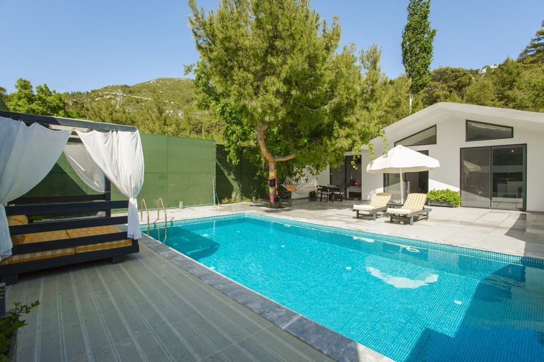 Beyaz villa, önünde mavi yüzme havuzu, şezlonglar, güneş şemsiyesi ve perdeli bir oturma alanı bulunuyor.