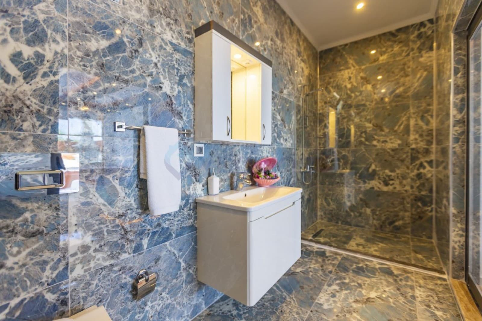 Mavi mermer kaplı duvarları olan banyoda beyaz lavabo, ayna ve duşakabin yer alıyor.