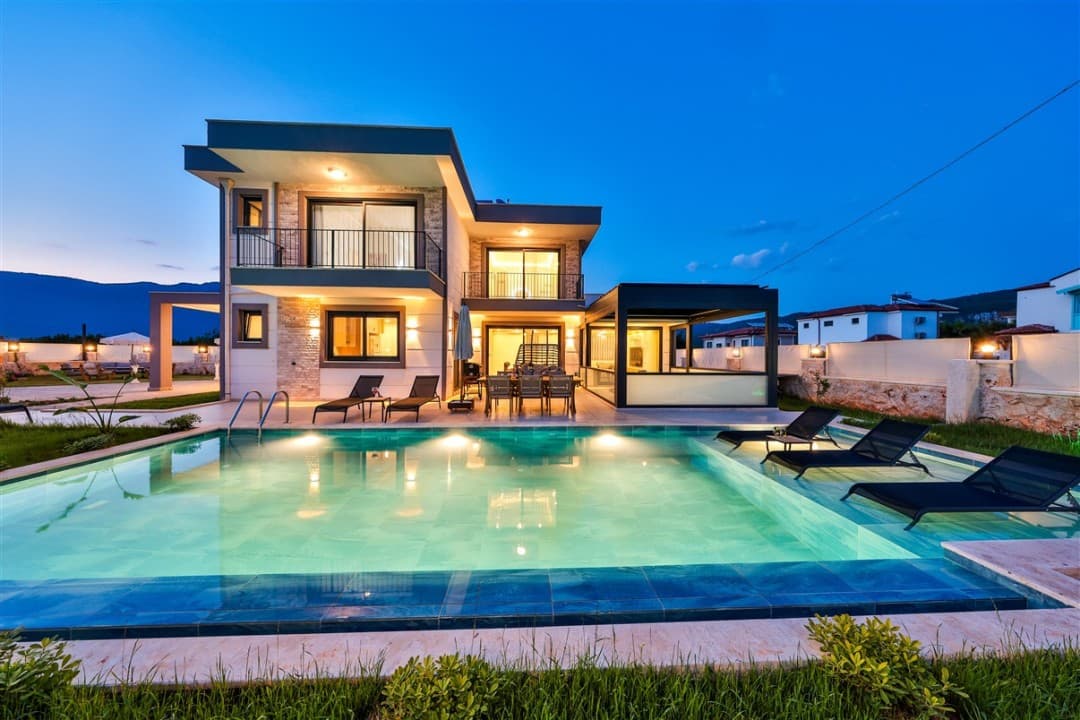 Alacakaranlıkta havuzu aydınlatılmış iki katlı modern villa ve şezlonglar.