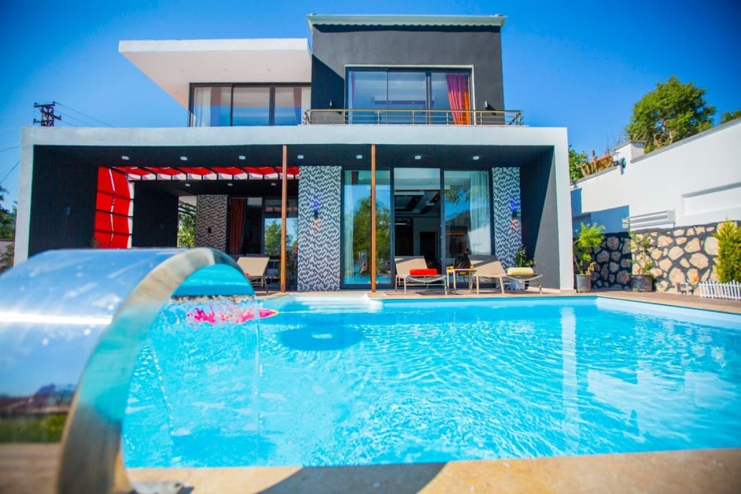 Açık havuzlu modern villanın önünde şezlonglar ve havuz suyu düşüren krom şelale yer alıyor.