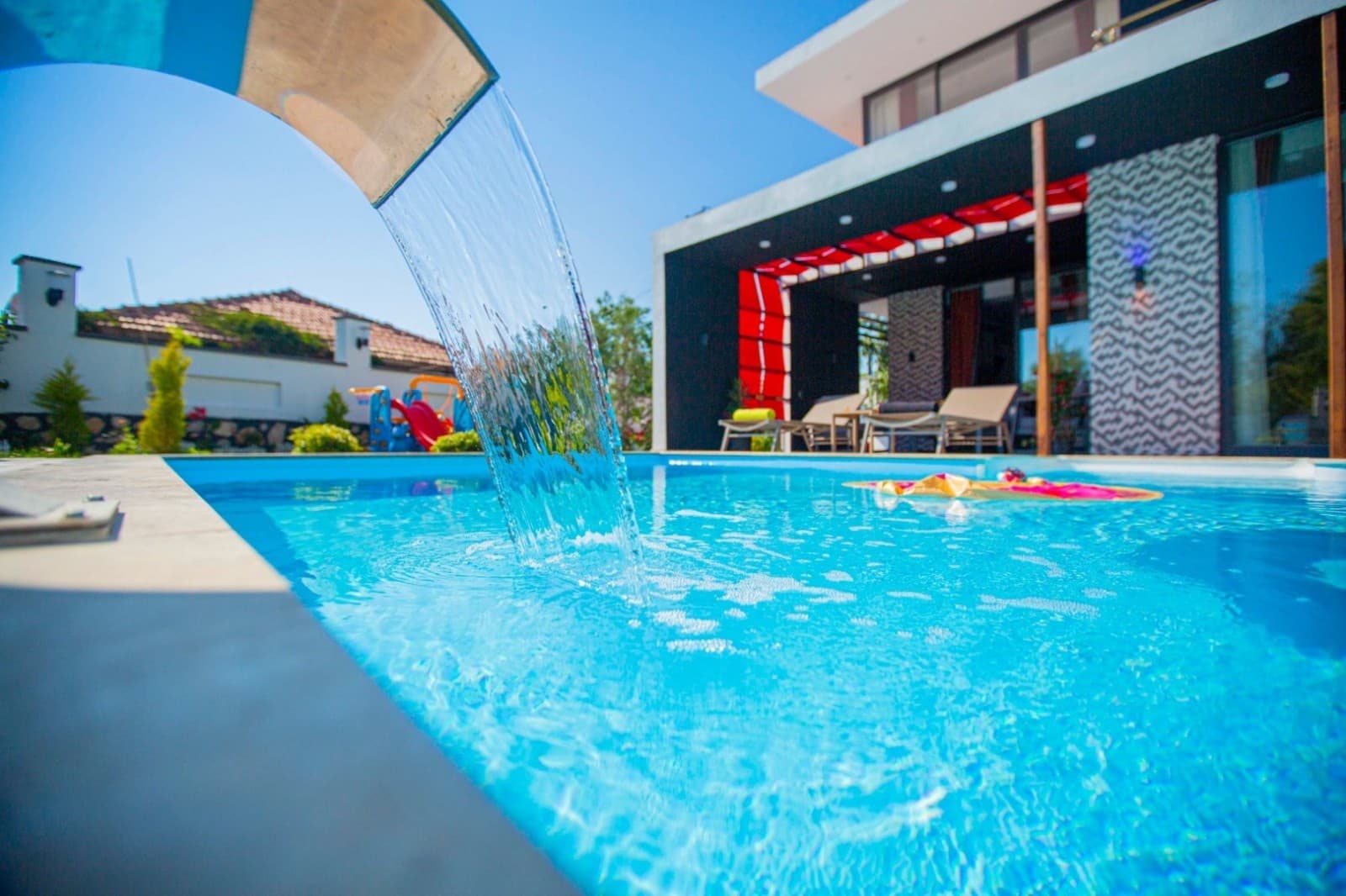 Havuz kenarında su şelalesi, şezlonglar ve arka planda beyaz villa.