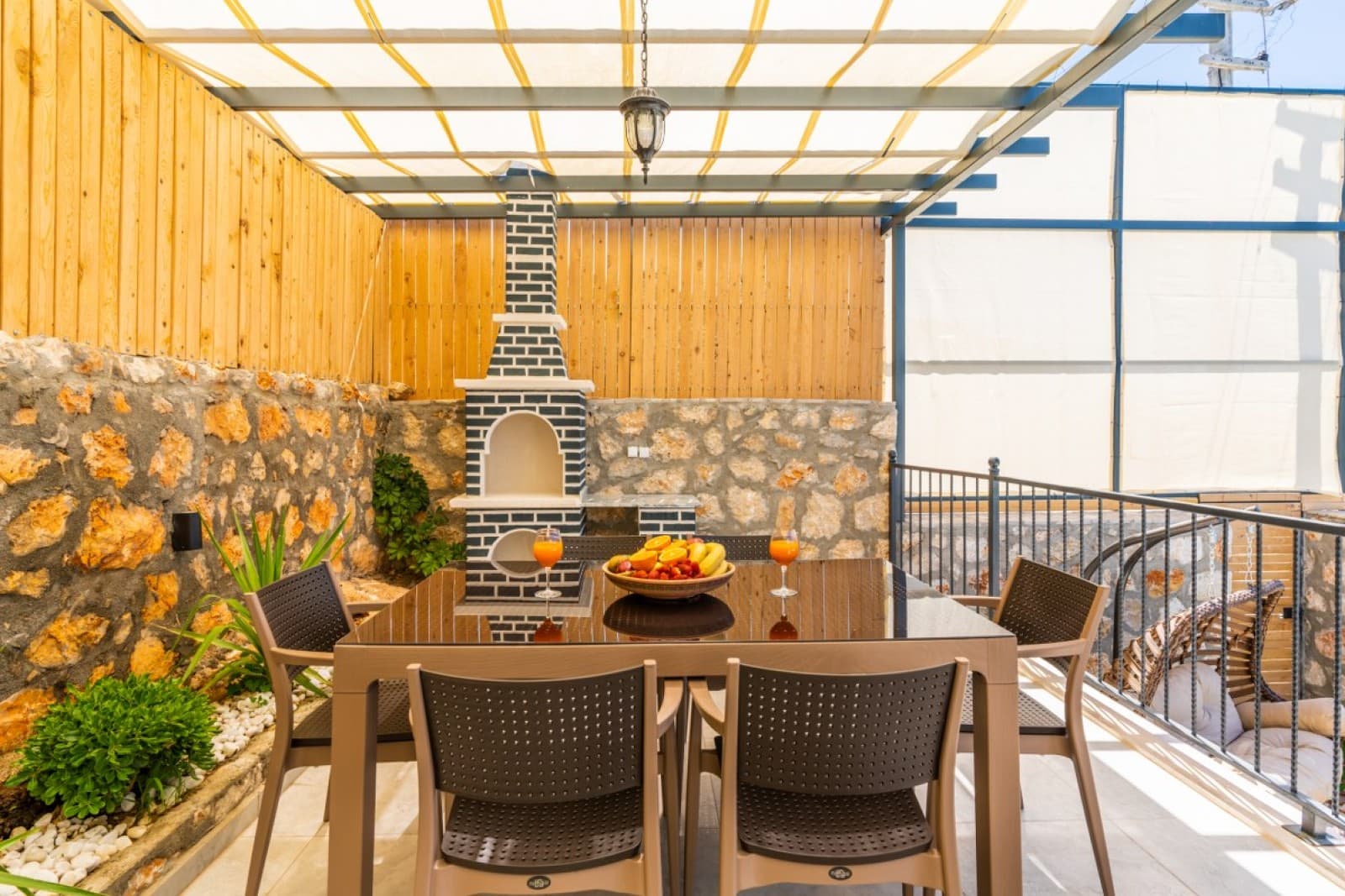Tente altındaki taş barbekü önünde cam yemek masası ve sandalyeler.