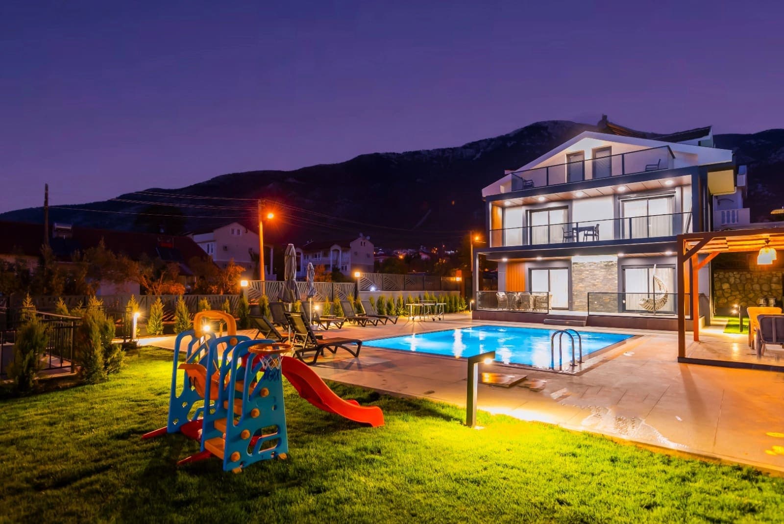 Akşam ışıklandırmalı havuz kenarında çocuk kaydırağı ve arkada villa dış cephesi.